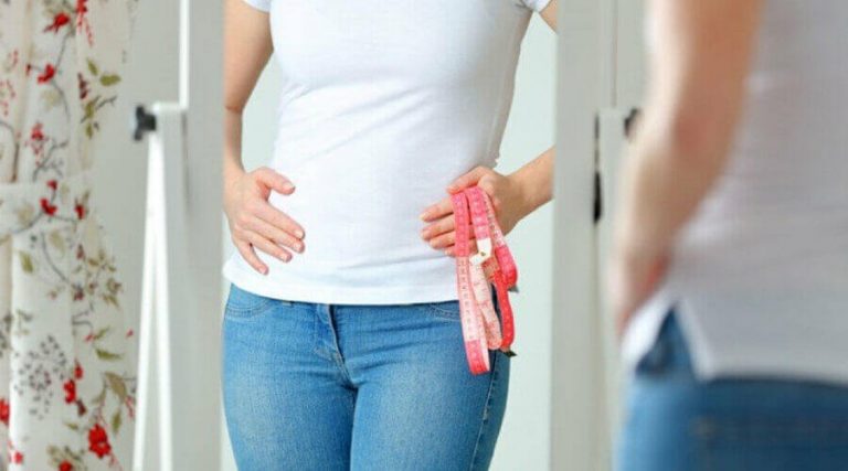 A girl is holding a measuring tape in her hand and looking at her stomach, feeling it appears fat.