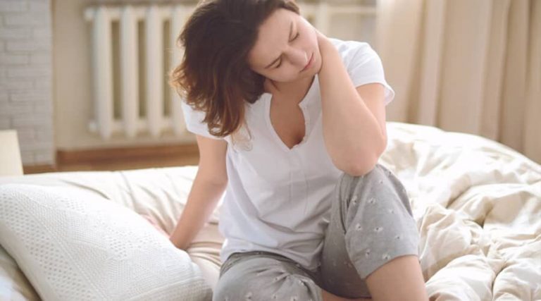 A girl sitting on her bed, holding her neck in pain after sleeping with the wrong pillow position.