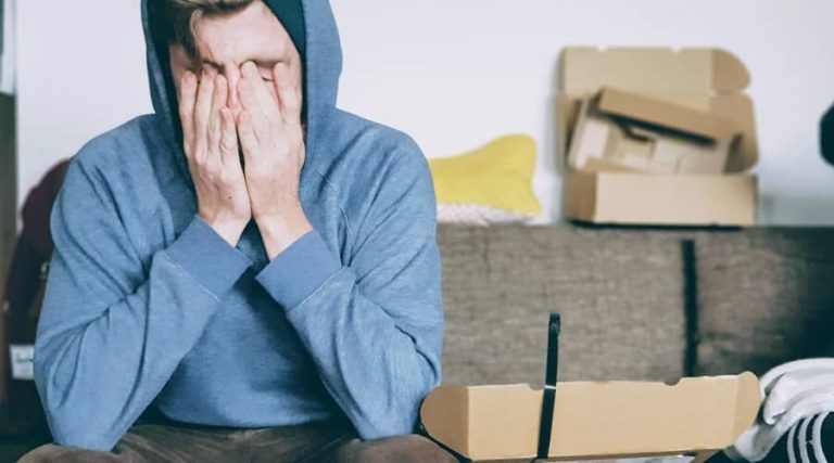 man wearing a hoodie sits with his hands covering his face, surrounded by cardboard boxes.