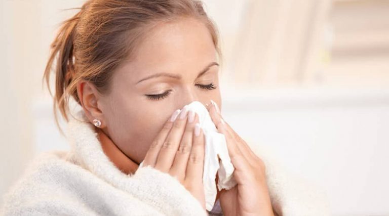 A woman wrapped in a blanket blows her nose with a tissue.