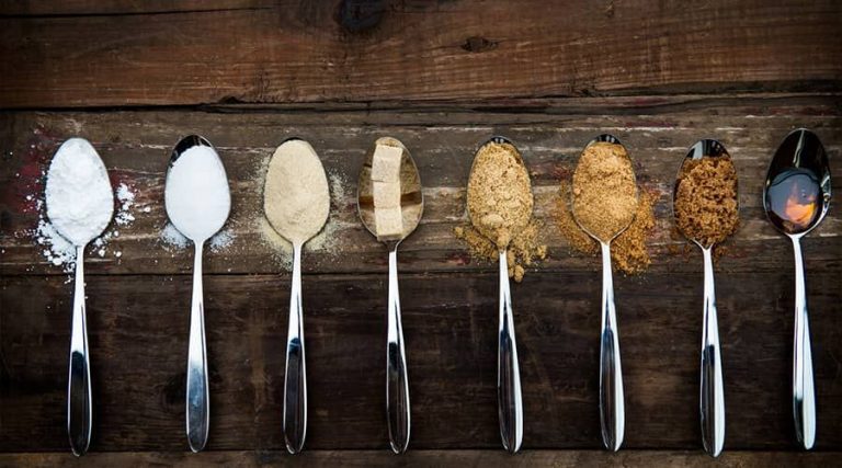 A row of spoons on a wooden surface, each holding a different type of sugar or sweetener, ranging from white powdered sugar to brown sugar and syrup