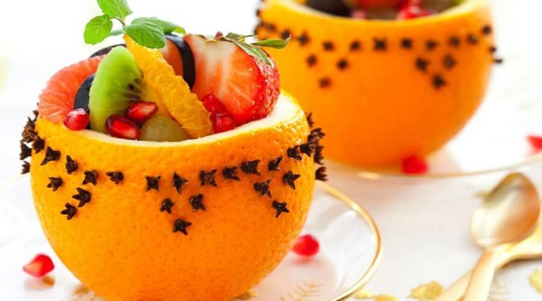 Decorative orange bowls filled with fresh fruit slices, garnished with cloves and a sprig of mint, placed on a white plate with a golden spoon nearby.