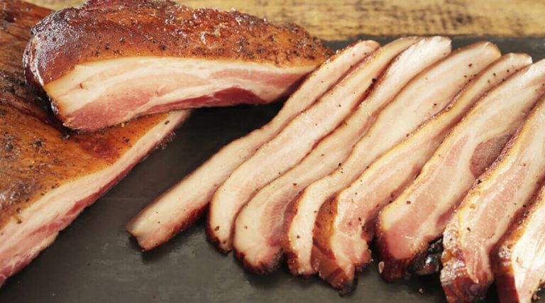 Close-up of thick slices of smoked bacon arranged on a dark cutting board, showcasing its marbled texture and seasoning.