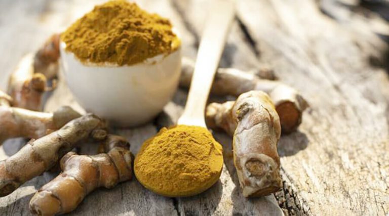 A close up of turmeric powder in a bowl and spoon surrounded by fresh turmeric roots on a rustic wooden surface