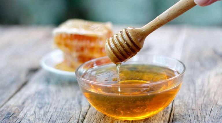 A clear glass bowl filled with golden honey, with a wooden honey dipper drizzling honey above it.