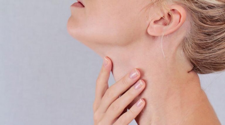 A close-up of a woman gently touching her throat with her hand, suggesting discomfort or pain. Her head is slightly tilted back, highlighting the neck area.