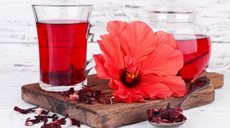 A glass of vibrant red hibiscus tea is placed on a wooden board, accompanied by a teapot filled with the same tea