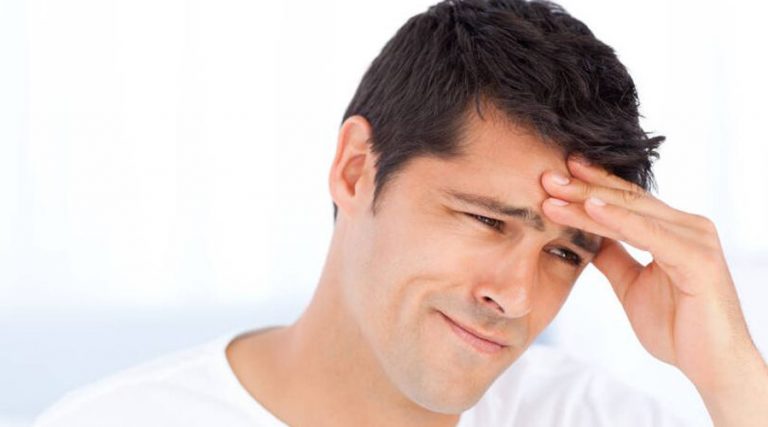 A close-up of a young man with dark hair, pressing his hand against his forehead with a pained or stressed expression.