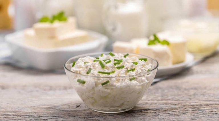 a bowl of grated cottage cheese. In the background, a plate holds a block of cottage cheese on a rustic wooden table.
