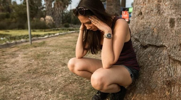 A young woman with long brown hair sits on the ground, leaning against a tree trunk in an outdoor park setting.