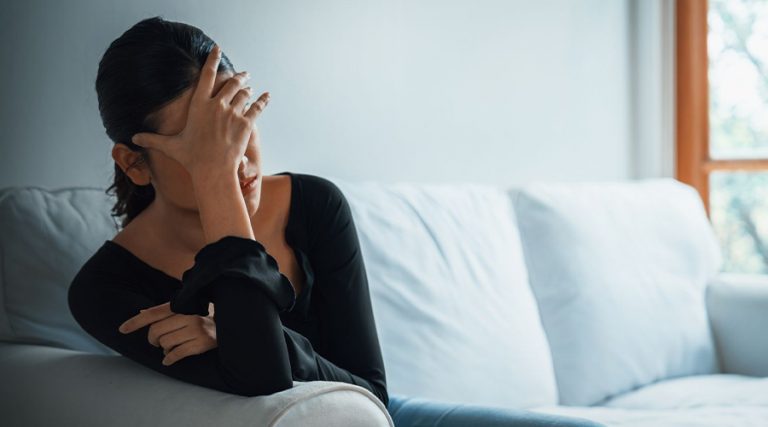 A woman with dark hair tied back, wearing a black long-sleeved top, sits on a light-colored couch with her hand covering her face.