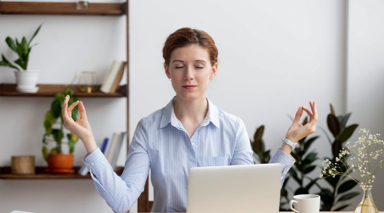 A woman with red hair, wearing a light blue striped shirt, sits at her desk with her eyes closed, meditating.