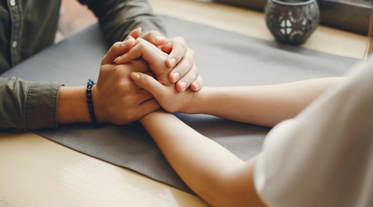 Two people are sitting at a table, holding hands gently in a supportive and comforting manner.