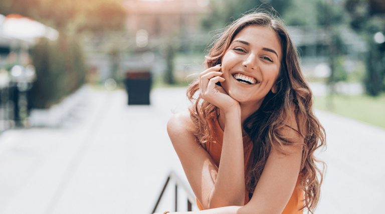 A young woman with long, wavy hair is smiling brightly while posing outdoors.