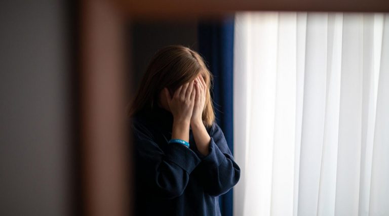 A woman with shoulder-length hair stands near a window with white curtains, covering her face with both hands