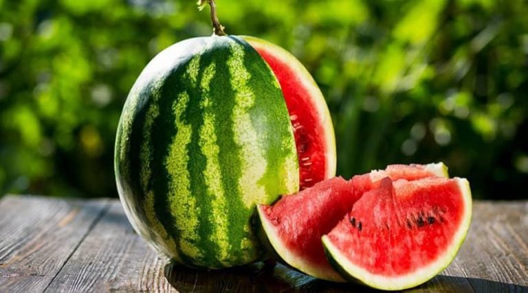 A whole watermelon with a deep green rind and vibrant red flesh is placed on a rustic wooden table outdoors.