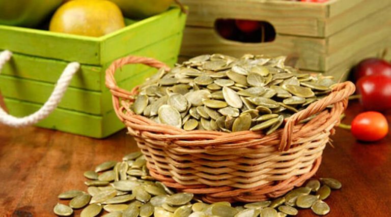 A woven basket filled with green pumpkin seeds spilling onto a wooden table, with green and wooden crates containing fruits in the background.