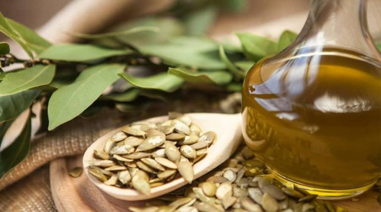 A wooden spoon filled with pumpkin seeds is placed on a natural wooden surface, accompanied by a glass bottle of golden pumpkin seed oil.