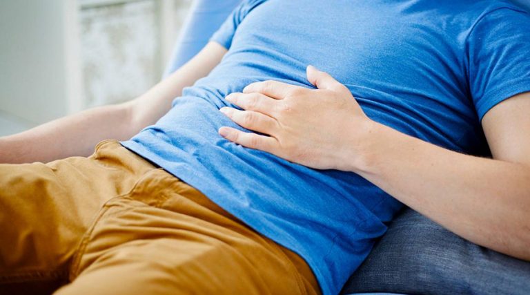A man wearing a blue t-shirt and mustard-colored pants is lying on a couch with his hand placed on his stomach, indicating discomfort or pain.