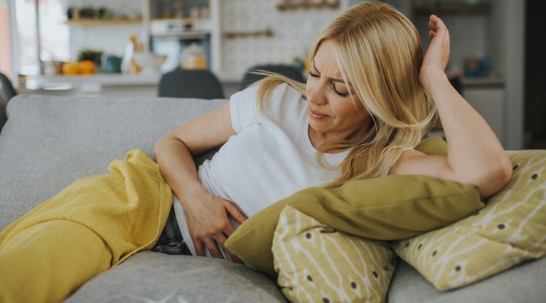 A blonde woman wearing a white t-shirt lies on a gray couch, clutching her stomach with a pained expression on her face.