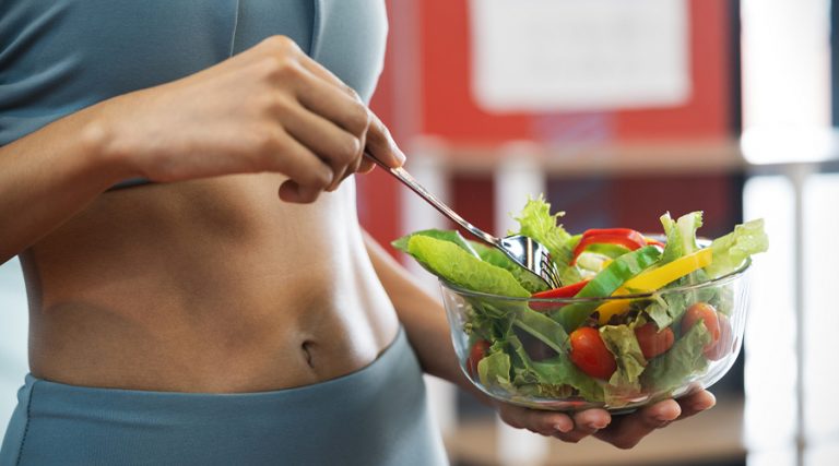 A woman in athletic wear holds a glass bowl filled with a colorful salad, including cherry tomatoes, lettuce, sliced bell peppers, and cucumbers.