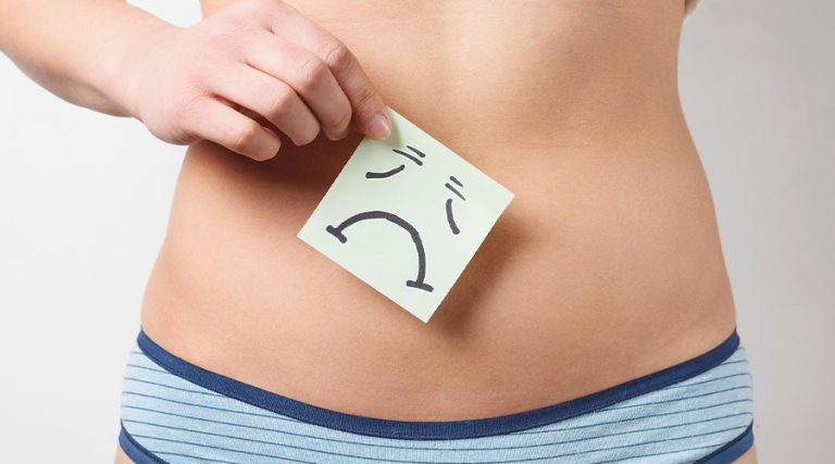 A person holding a light green sticky note with a sad face drawing over their bare stomach, symbolizing abdominal discomfort or digestive issues.