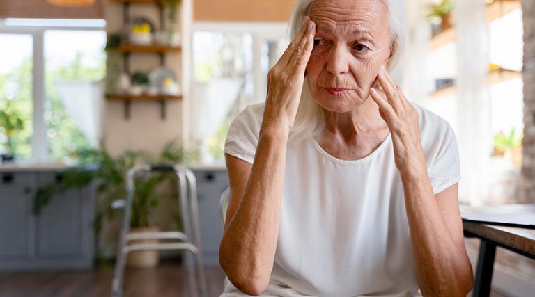 An elderly woman with light skin and white hair is seated in a well-lit kitchen, pressing her fingers to her temples with a concerned and pensive expression