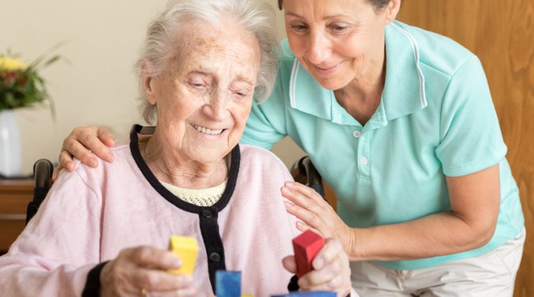 An elderly woman with light skin, seated and wearing a pink cardigan, is engaging with colorful building blocks as part of a cognitive activity