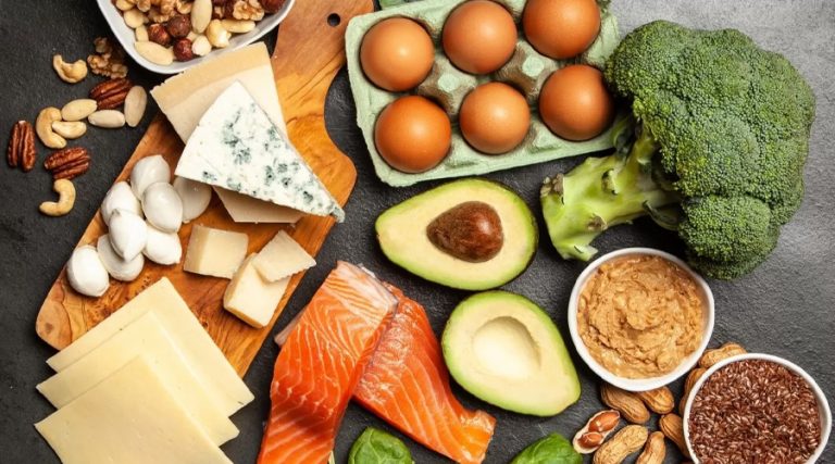 A collection of keto-friendly foods displayed on a dark surface, including salmon, eggs, avocado, broccoli, assorted cheeses, nuts, seeds, peanut butter, and spinach