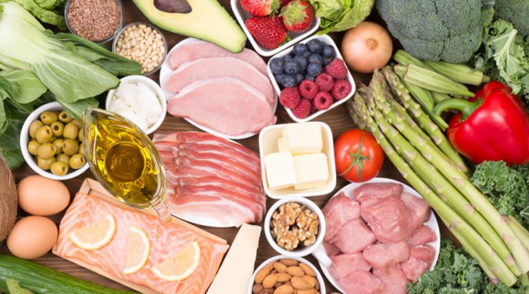 A variety of low-carb, nutrient-rich foods arranged on a wooden surface, including salmon, chicken, pork, eggs, leafy greens, asparagus, avocado, nuts, seeds, berries, olives, butter, and olive oil.