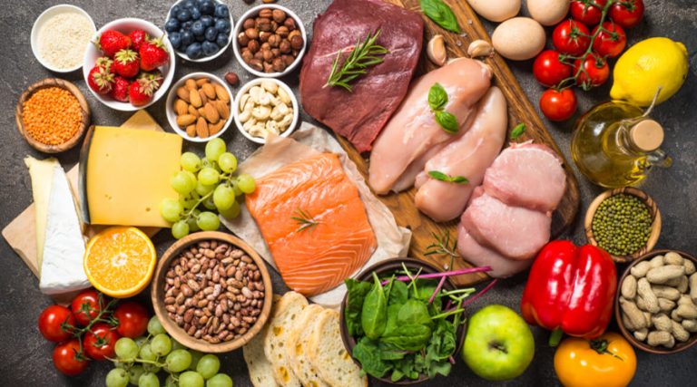 A diverse assortment of whole food ingredients arranged on a dark surface, including fresh fruits, vegetables, lean meats, fish, nuts, seeds, legumes, cheese, eggs, and bread.