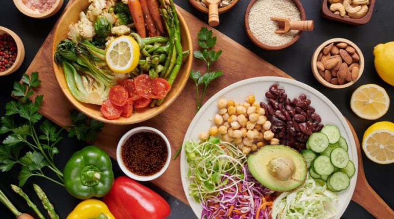 A vibrant plant-based vegan meal displayed with a plate of fresh vegetables, including avocado, cucumber, sprouts, cabbage, chickpeas, and kidney beans, alongside a bowl of roasted vegetables such as carrots, Brussels sprouts, and asparagus.