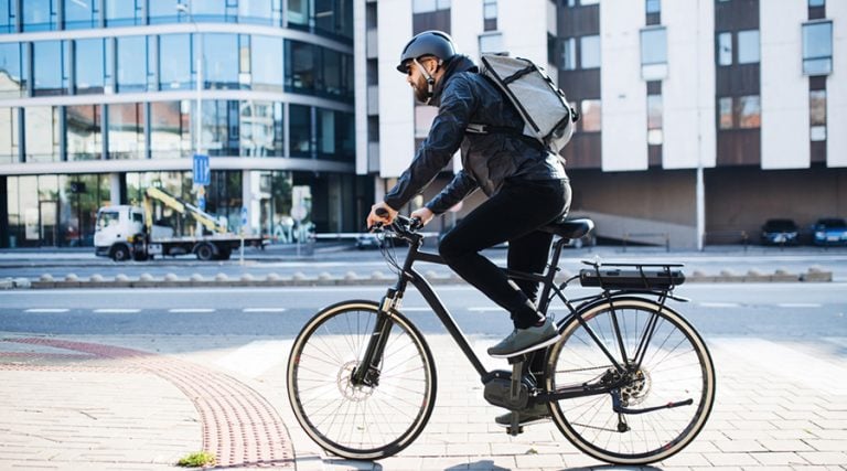A man wearing a helmet, sunglasses, and a backpack is riding an electric bicycle through an urban setting with modern glass buildings and a truck in the background