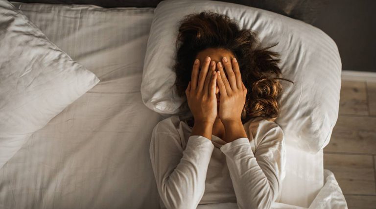 A woman with curly hair lies in bed wearing a white long-sleeved shirt, covering her face with both hands