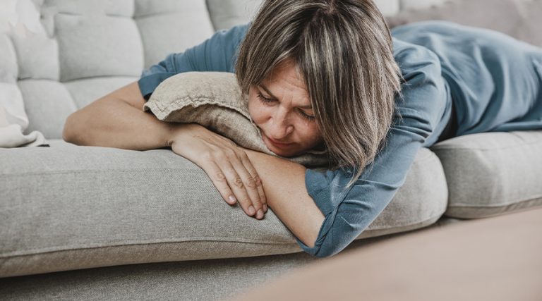 A woman in a blue dress lies face down on a gray couch, resting her head on a beige cushion