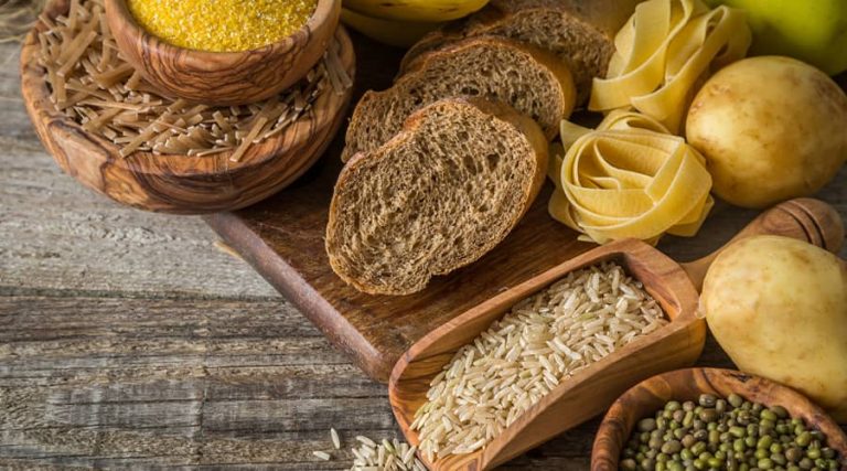 A variety of carbohydrate-rich foods displayed on a rustic wooden surface, including whole grain bread slices, uncooked rice in a wooden scoop, pasta nests, potatoes, green mung beans, and different grains such as bulgur and brown rice in wooden bowls.